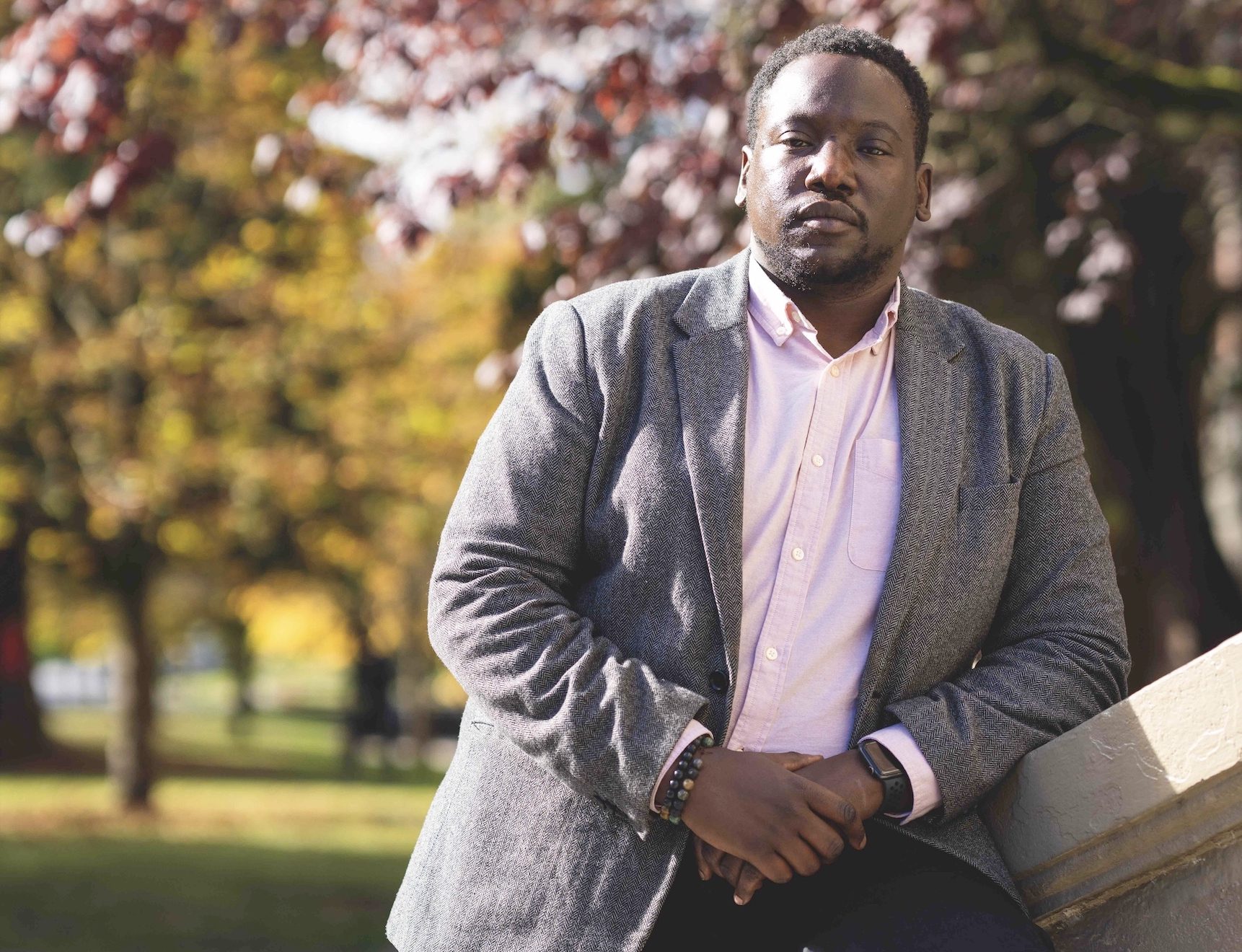 A Black man in a grey blazer leans against a railing.